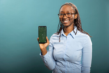 Cheerful black female model holds mobile device displaying green screen, posing against blue background with friendly smile. African American woman holding smartphone with blank mockup template.
