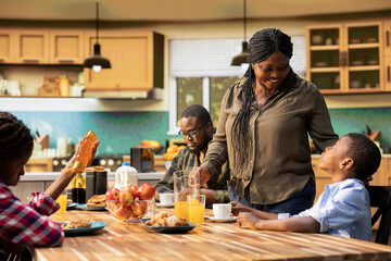 Black mother serving breakfast at the table and pouring orange juice in a glass, young family enjoying eggs and bacon together. Morning routine filled with joy and love shared over meal.