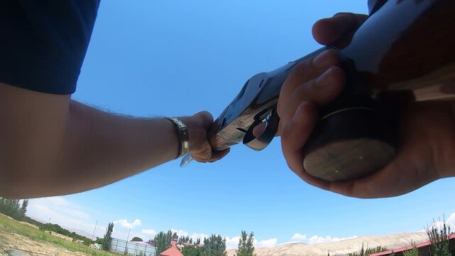 Action camera POV of a sportsman loading and firing a shotgun at a shooting range. The footage captures the weapon recoil against a clear blue sky and mountains.

