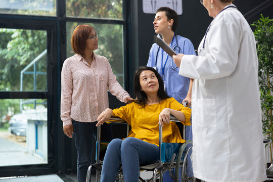 Medic chatting with asian woman wheelchair user about managing chronic condition during checkup visit. Medical expert offering physical support and lifestyle recommendations to improve mobility.