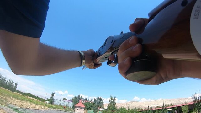 Action camera POV of a sportsman loading and firing a shotgun at a shooting range. The footage captures the weapon recoil against a clear blue sky and mountains.

