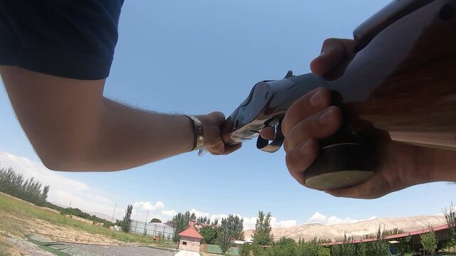 Action camera POV of a sportsman loading and firing a shotgun at a shooting range. The footage captures the weapon recoil against a clear blue sky and mountains.

