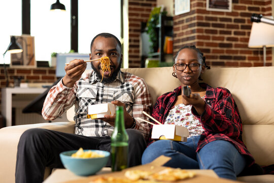 African american couple relaxing at home, sharing takeaway food on couch. Man uses chopsticks to eat noodles as woman flips through tv channels, enjoying cozy and casual evening together.