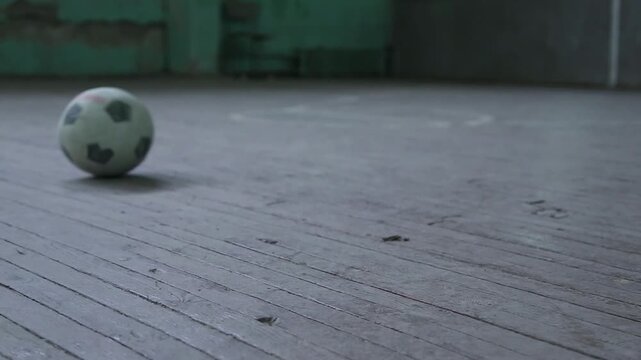 A deflated soccer ball spins slowly on worn wooden floorboards. In the background, a cracked teal wall suggests an old, perhaps abandoned, indoor gymnasium or sports hall.


