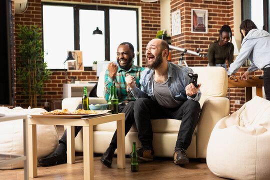 Black and white men seated on couch with fists raised in celebration after winning online gaming tournament. Multiethnic male friends cheering in excitement, enjoying fun weekend in apartment. - Powered by Adobe