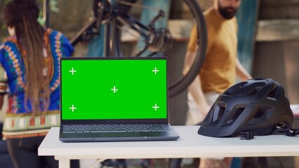 Close-up of laptop with greenscreen display placed near helmet while interracial couple performs bike maintenance in background. In yard minicomputer is on table showing isolated chromakey template.