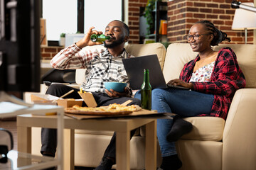 Smiling black woman works remotely on laptop while man enjoys beer beside her on couch. Young couple shares casual pizza night, watching TV and relaxing together in cozy brick wall apartment.