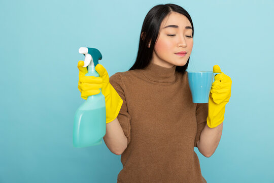 Female janitor pauses cleaning to enjoy coffee break, holding mug near mouth and spray bottle in hand. Asian woman looking at cup, having peaceful moment amid household chores.