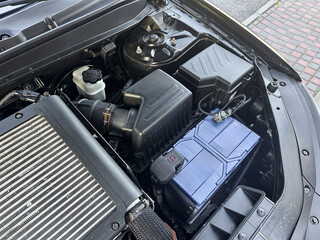 Close-up view of a car engine bay showcasing components like the battery, air intake, and cooling system during mid-afternoon in an urban environment