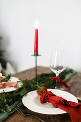 Merry Christmas! Stylish table setting for christmas family dinner. Modern plate and cutlery with napkin, glassware, green and red ribbons, fir branches and candle on rustic table