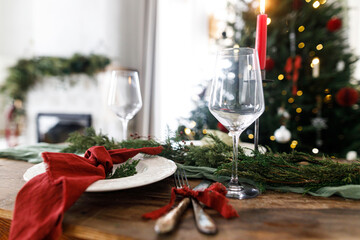 Merry Christmas! Stylish table setting for christmas family dinner. Modern plate and cutlery with napkin, glassware, green and red ribbons, fir branches and candle on rustic table at xmas tree