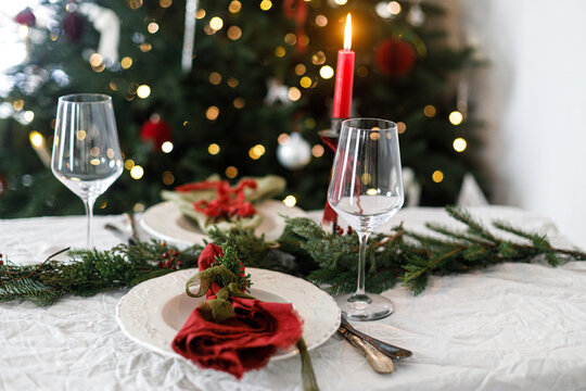 Merry Christmas! Stylish table setting for christmas family dinner. Modern plate and cutlery with napkin, glassware, green and red ribbons, fir branches and candle on rustic table at xmas tree