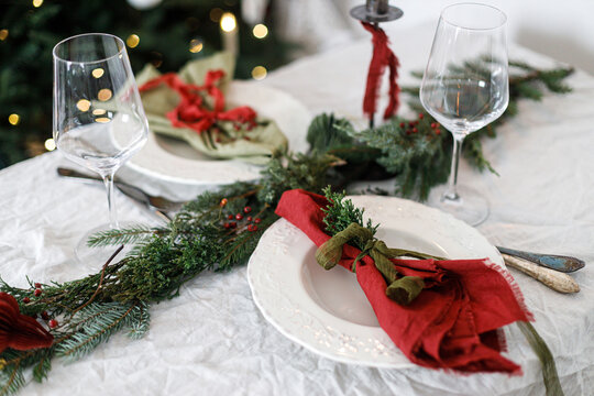 Merry Christmas! Stylish table setting for christmas family dinner. Modern plate and cutlery with napkin, glassware, green and red ribbons, fir branches on rustic table at xmas tree