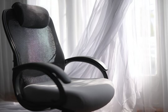 Black mesh office chair sits empty by bright window, white sheer curtains gently blowing.