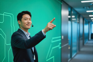 A professional Asian man in a suit wearing a headset points confidently at a futuristic green wall with circuit-like patterns, standing in an office hallway.