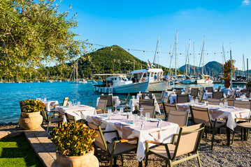 Traditional Greek restaurant tables with fishing boats in background in Nidri port before sunset, Lefkada island, Greece