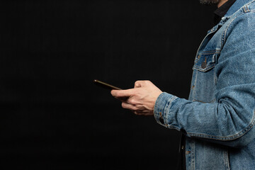 a man at the right side of the screen wearing a denim jacket texting with a smartphone and a black background