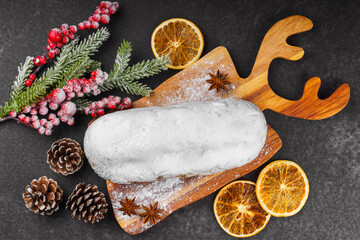 Classic Christmas Stollen covered with powdered sugar icing on wooden board with dried oranges, pine branches and cones on black textured surface, top view. Traditional holiday food, sweet fruit loaf