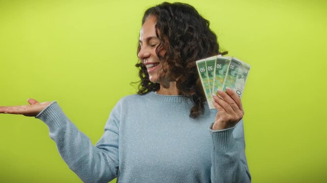 Middle-aged woman holding israeli shekel banknotes against a vibrant yellow background, smiling while extending her hand, conveying a sense of wealth and financial happiness.