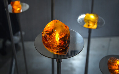 Large, deep orange amber gem glows on a small, dark metal pedestal. It is one of several similar pieces displayed on thin metal rods against a dark background