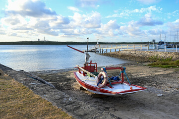 small fishing boat on dry land, potengi river natal brazil, local maritime scene traditional red and white boat, boat ramp and pier, cloudy sky landscape vessel on shore at low tide, coastal area