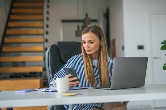 Woman working from home office using laptop and phone - Powered by Adobe