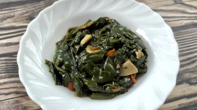 Stir-fried genjer vegetables are served on a white plate on a wooden table. Genjer vegetables, Limnocharis flava, Sawah-lettuce, or yellow velvetleaf, are aquatic plants that can be cooked into dishes