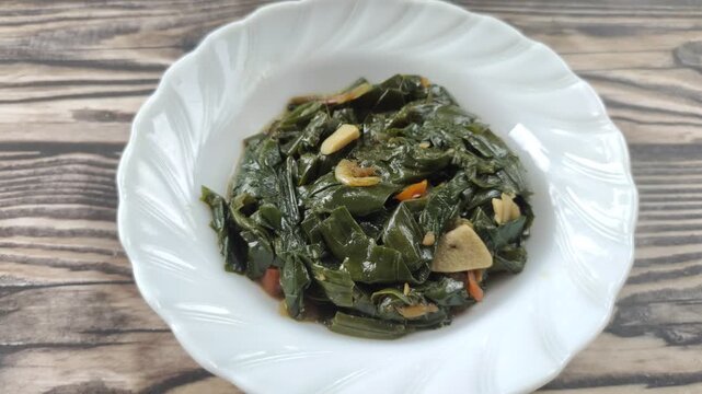 Stir-fried genjer vegetables are served on a white plate on a wooden table. Genjer vegetables, Limnocharis flava, Sawah-lettuce, or yellow velvetleaf, are aquatic plants that can be cooked into dishes