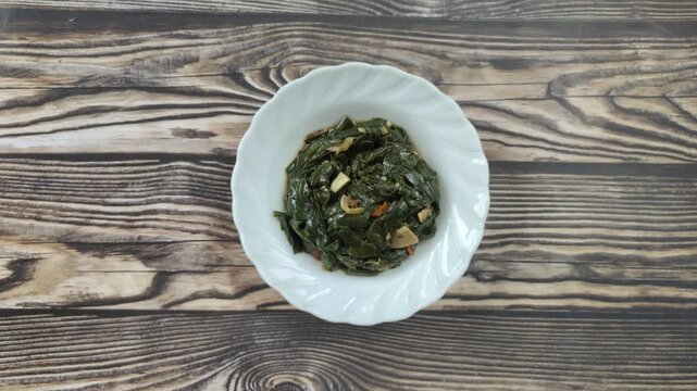 Stir-fried genjer vegetables are served on a white plate on a wooden table. Genjer vegetables, Limnocharis flava, Sawah-lettuce, or yellow velvetleaf, are aquatic plants that can be cooked into dishes