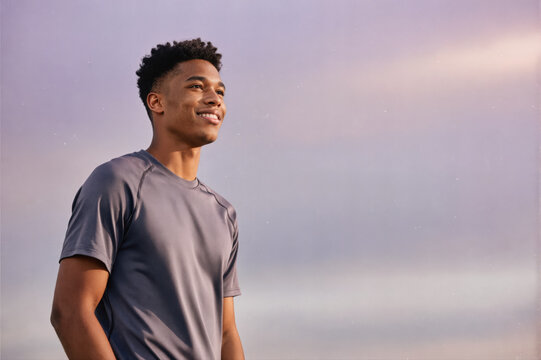A young man in a gray athletic shirt smiling and looking to the side against a soft purple sky, captured in a lifestyle portrait. - Powered by Adobe