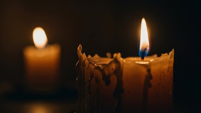 Two lit candles stand in the darkness the closer one featuring drips of wax down its side