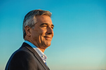 A side profile portrait of a smiling middle-aged man with gray hair wearing a suit and tie against a clear blue sky.