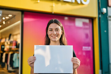 A young woman with long brown hair stands smiling in front of a brightly colored storefront, holding a blank white signboard in a vibrant urban setting.