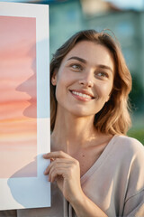 A smiling young woman holding a large art print with a sunset design, photographed outdoors in natural light.