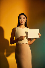 A smiling young Asian woman in a cream sleeveless top and beige skirt holding a blank white card, pointing at it with her index finger against a warm yellow and teal gradient background.