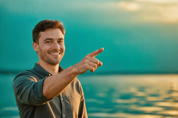 A smiling man pointing to the side while standing outdoors near a body of water during sunset, captured in a vibrant photo.