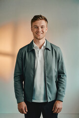 A smiling young man in a gray jacket and white shirt standing against a soft gradient background.