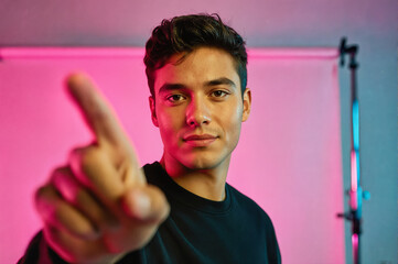 A young man in a black shirt pointing directly at the camera with his index finger, illuminated by vibrant pink and blue neon lighting against a studio backdrop.