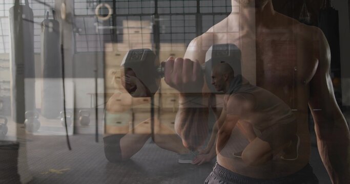 Holding hex dumbbell, shirtless man curling forearm at warehouse gym, with plyo boxes and rings