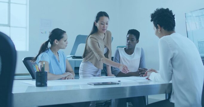 Pointing standing woman in tan blazer leaning over conference table in office showing plans, tablet