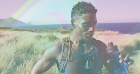 Standing hiker resting at coastal dune, holding blue water bottle, wearing dark tank top backpack
