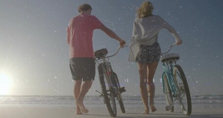 Naklejka premium Walking couple wearing cover-up and pink tee pushing two bicycles on wet sand, low sun reflecting