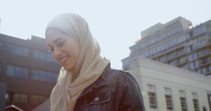 Smiling woman in light beige hijab and blue denim jacket checking phone on sunlit street, buildings - Powered by Adobe