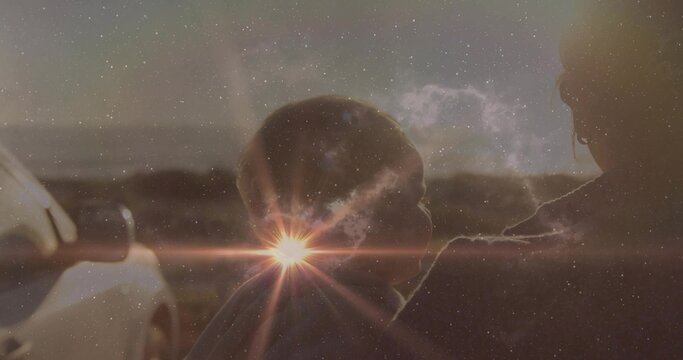 Fototapeta Cradling mother wearing coat and glasses holding hooded baby road under sun flare with parked car