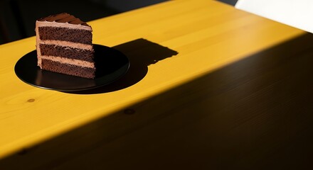 A delicious slice of layered chocolate cake with frosting on a black plate, sitting on a bright yellow wooden table with strong sunlight and shadows.