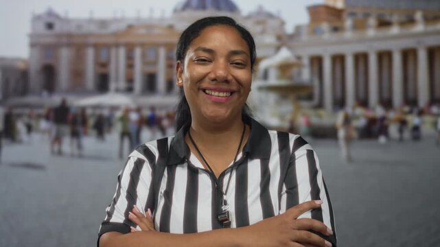 Woman referee smiles while folding arms before a historic church building in vatican square; confidence.