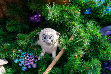 A small light colored toy monkey with long legs, big eyes, and small ears. The monkey character is hiding on a lush fir tree branch of a Christmas tree. There are purple and blue plastic decorations.