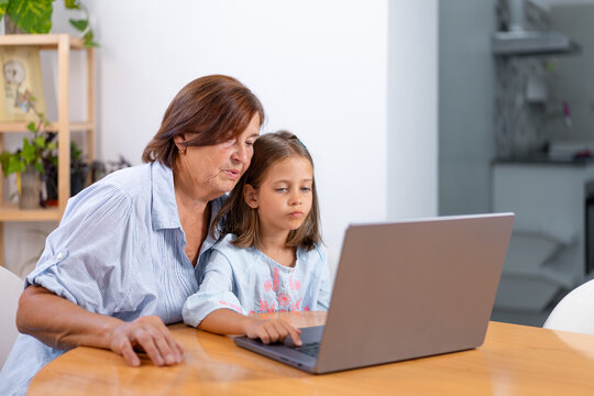 Grandmother and young girl sitting together at table focused on laptop screen sharing technology, learning, and quality family time at home