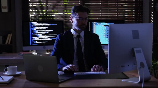 Programmer in suit and glasses working on computer and laptop at wooden table in office at night