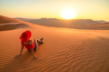 Woman sitting on the sand in the Empty Quarter desert of Oman at sunrise, enjoying the dawn view of the sand dunes. wearing red clothes that contrast with the golden sand.
